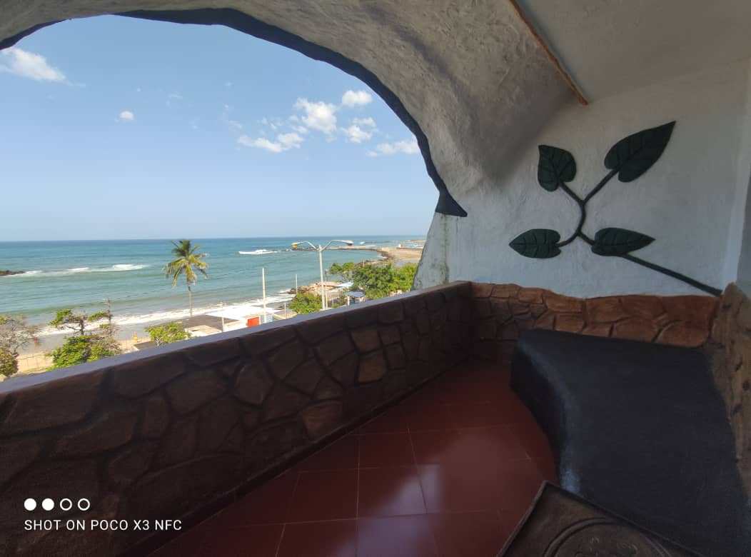 An outdoor balcony area presents a view of the Caribbean Sea, with waves gently lapping against the shore. The curved wall features decorative leaf motifs, and a stone seating area offers a place to relax. Clear skies and palm trees are visible in the distance.