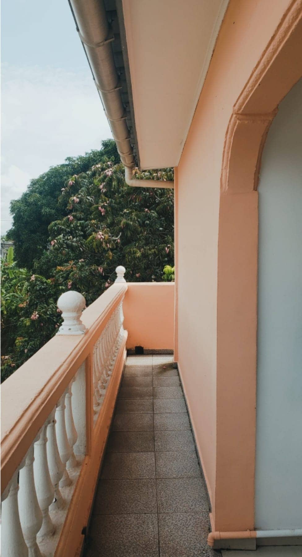 A narrow balcony is presented, featuring a pink stucco wall and a white railing. The tiled floor leads to a view of lush green trees just beyond the railing, creating a sense of seclusion and connection to nature.