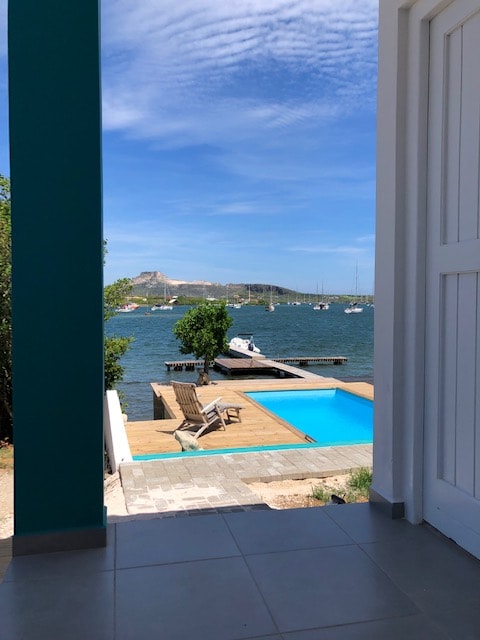 A view from the entrance showcases a serene swimming pool and a wooden dock extending into the water. Lush greenery and a mountain backdrop enhance the scene, while a sunny sky with scattered clouds adds brightness to the tranquil setting.