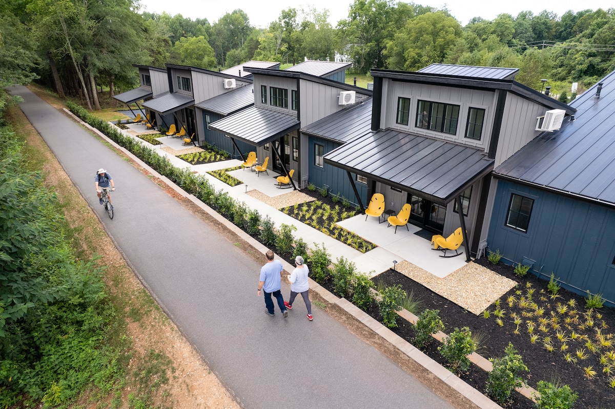 The exterior of the Wedge Cottages is showcased along a paved pathway, with bright yellow chairs arranged on porches. Lush landscaping with shrubs and gravel accents enhances the appeal. A cyclist is visible on the pathway, illustrating the connection to the Swamp Rabbit Trail.