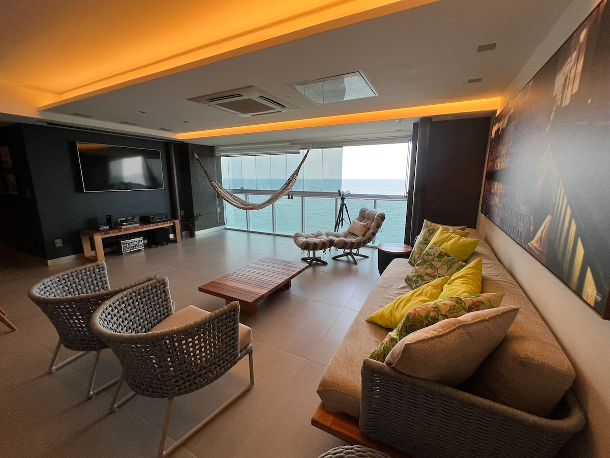 The living area features a comfortable sofa adorned with colorful cushions, accompanied by two woven chairs. A wooden coffee table sits centrally, while soft lighting enhances the space. Large glass windows provide a clear view of the ocean, with a hammock suspended nearby.