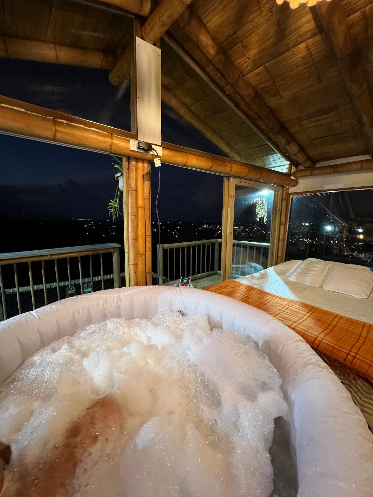 A bubble-filled jacuzzi is positioned beside a spacious bed, with a soft blanket draped casually over it. Large windows offer an expansive view of the night sky and the surrounding scenery, creating a serene atmosphere in a natural setting.
