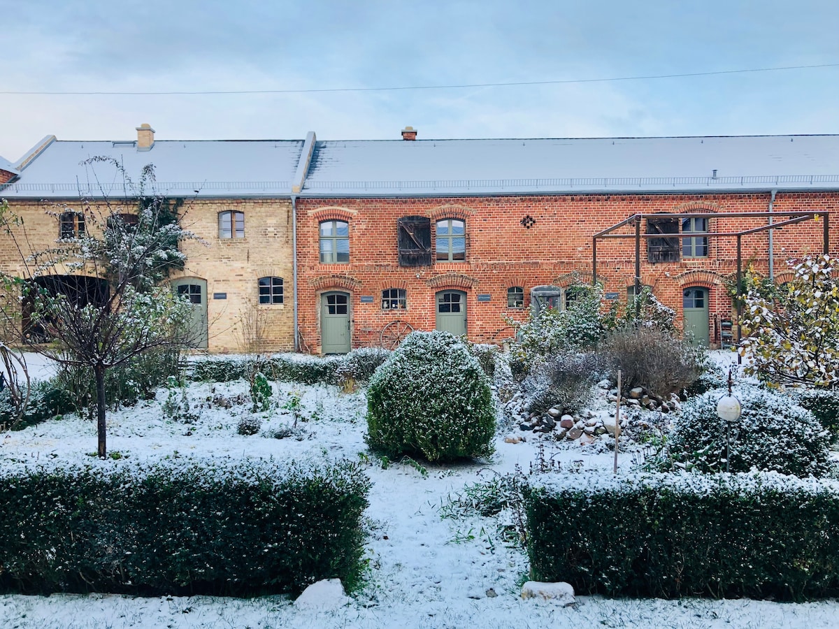 The courtyard of a historic farm is covered in a light layer of snow. Brick buildings with green doors are visible in the background. Neatly trimmed hedges and garden beds are outlined by the snow, enhancing the serene winter landscape.