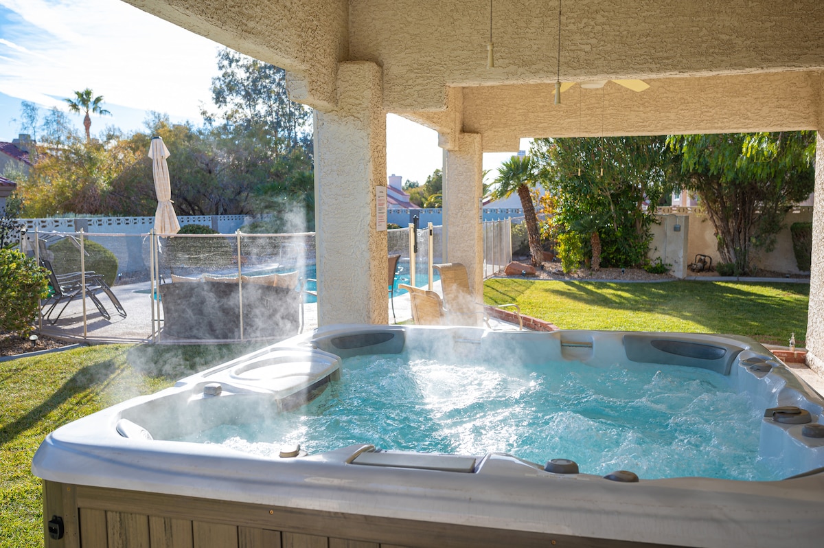 A hot tub spa is positioned under a covered patio, with steam rising from the water's surface. Lush green grass surrounds the spa, while a pool and lounge chairs are visible in the background, framed by palm trees and a serene backyard setting.