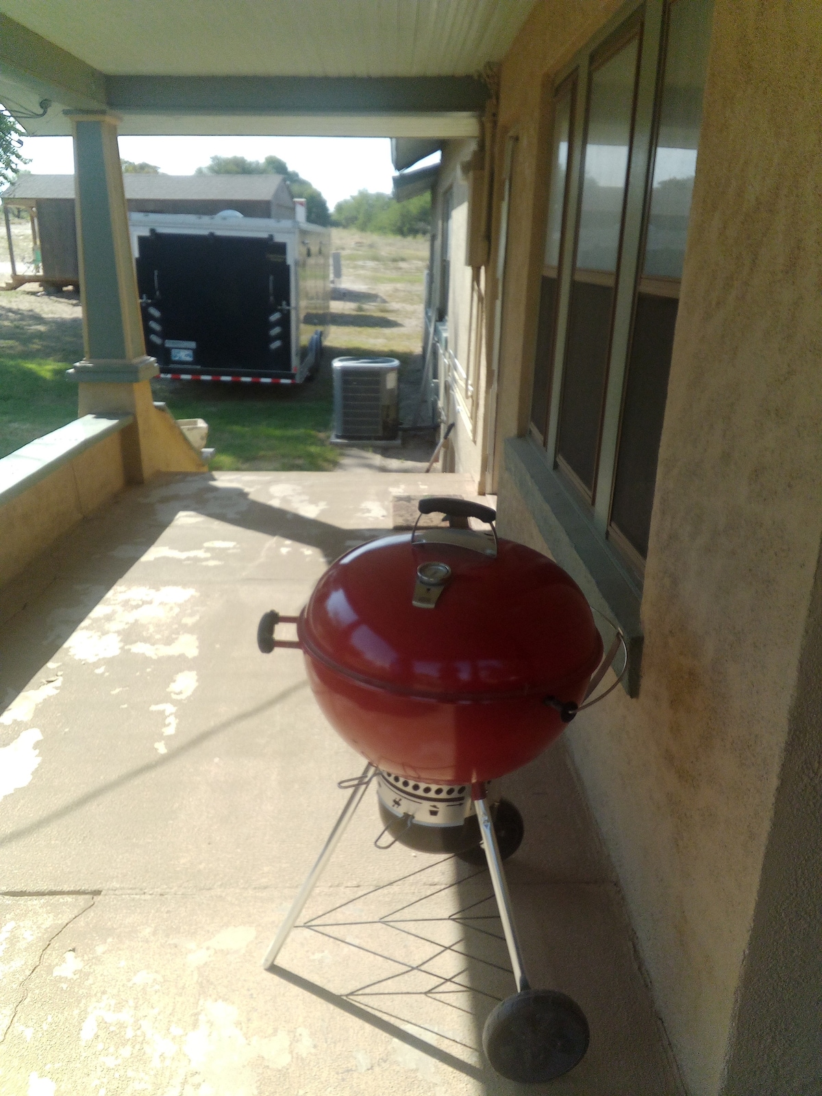 A red barbecue grill is positioned on a concrete patio, adjacent to the house. The background features an enclosed trailer and an air conditioning unit. Natural surroundings are visible, providing an open and spacious ambiance.