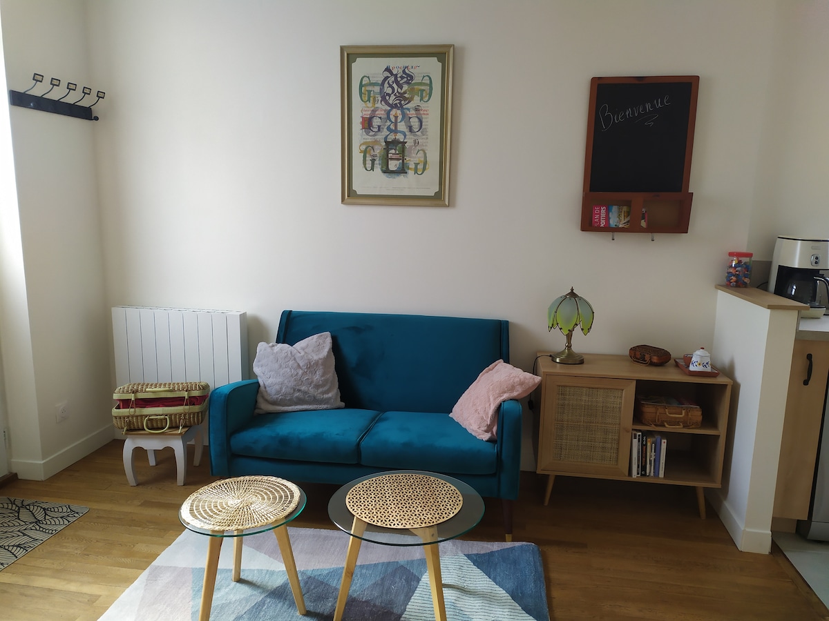 A cozy living area is featured, furnished with a teal sofa adorned with two cushions. A decorative lamp illuminates the space, while two round coffee tables are positioned on a layered rug. A framed artwork hangs on the wall, and a chalkboard with welcoming text is visible.