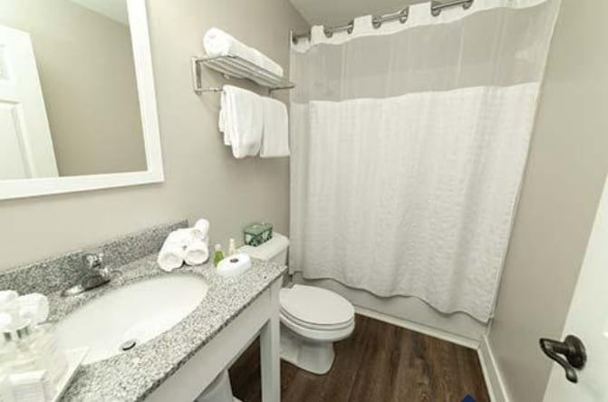 A compact bathroom space is equipped with a granite countertop sink, complemented by neatly arranged towels on a shelf above. A white shower curtain creates a clean look, with light-colored walls providing a neutral backdrop. A modern toilet is positioned adjacent to the sink area.