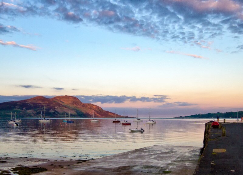 A serene waterfront scene shows a calm bay with several boats moored. Gentle ripples reflect the soft hues of the sky at dusk, while a distant hill provides a natural backdrop. The waterway is bordered by a simple stone pier, adding to the tranquil atmosphere.