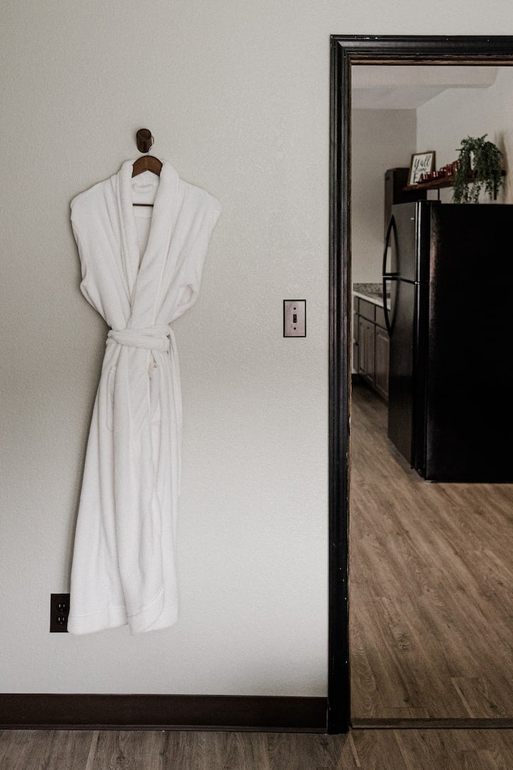 A plush white robe is hung on a wall hook, indicating comfort and relaxation. A doorway opens to the kitchen area, featuring a glimpse of a black refrigerator and countertops, with warm wooden flooring leading throughout the space.