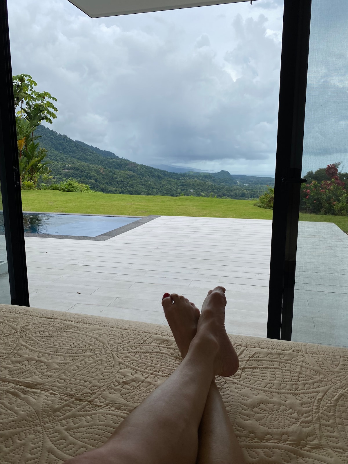 A tranquil indoor space offers a view of the expansive outdoor area featuring a sleek infinity pool and lush greenery. The floor-to-ceiling glass doors provide an unobstructed perspective of the mountains and ocean beyond, enhancing the natural light throughout the room.