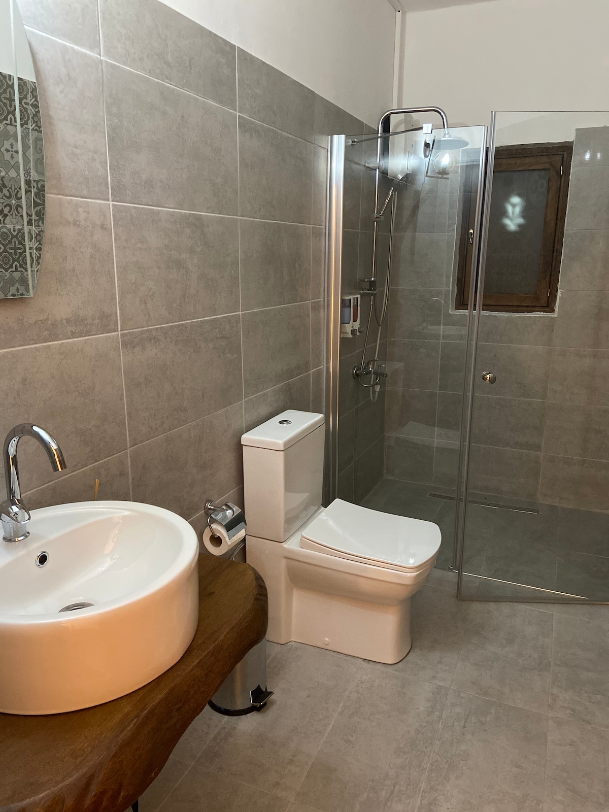 The bathroom features a modern design with neutral-toned tiles. A spacious, circular sink rests on a wooden countertop, while a glass-enclosed shower is equipped with a showerhead and wall-mounted controls. A window allows natural light, enhancing the overall brightness of the space.