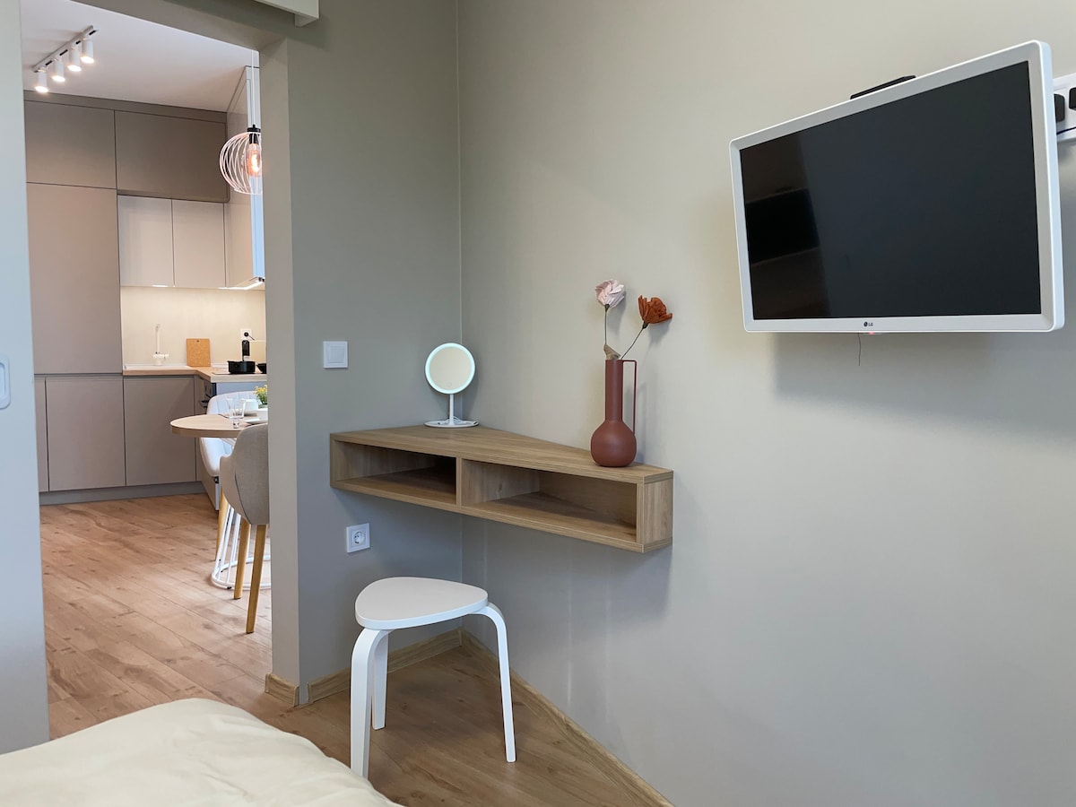 A modern wall-mounted TV is positioned above a wooden desk, featuring a small white chair. A vase with flowers and a round mirror are neatly arranged on the desk. In the background, a well-equipped kitchen is visible, showcasing minimalist cabinetry.