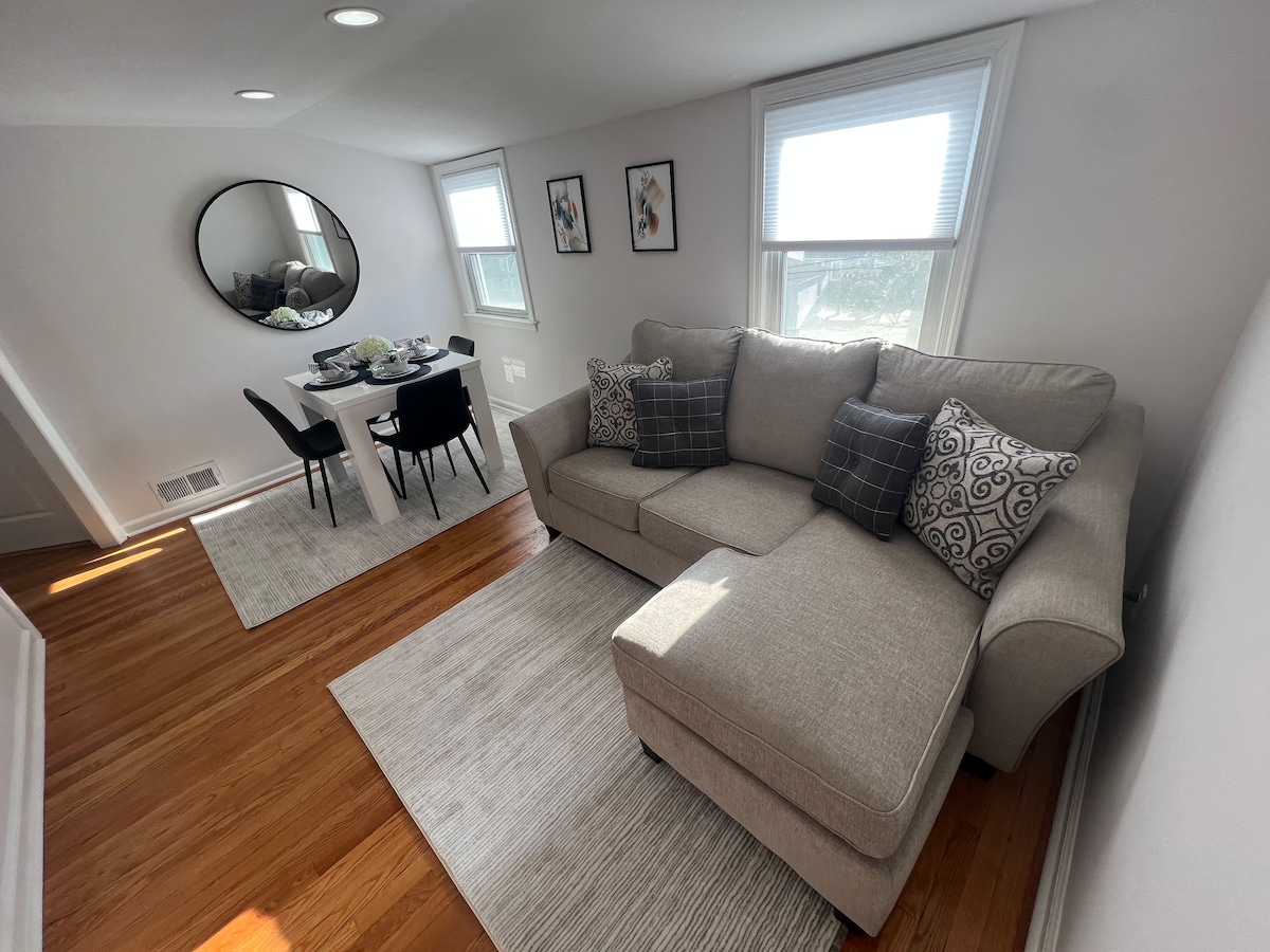The living area features a spacious sectional sofa adorned with decorative pillows, positioned opposite a dining table set for four. Natural light enters through two windows, enhancing the open atmosphere. Soft cream rugs complement the hardwood floor, providing a cozy and welcoming space.