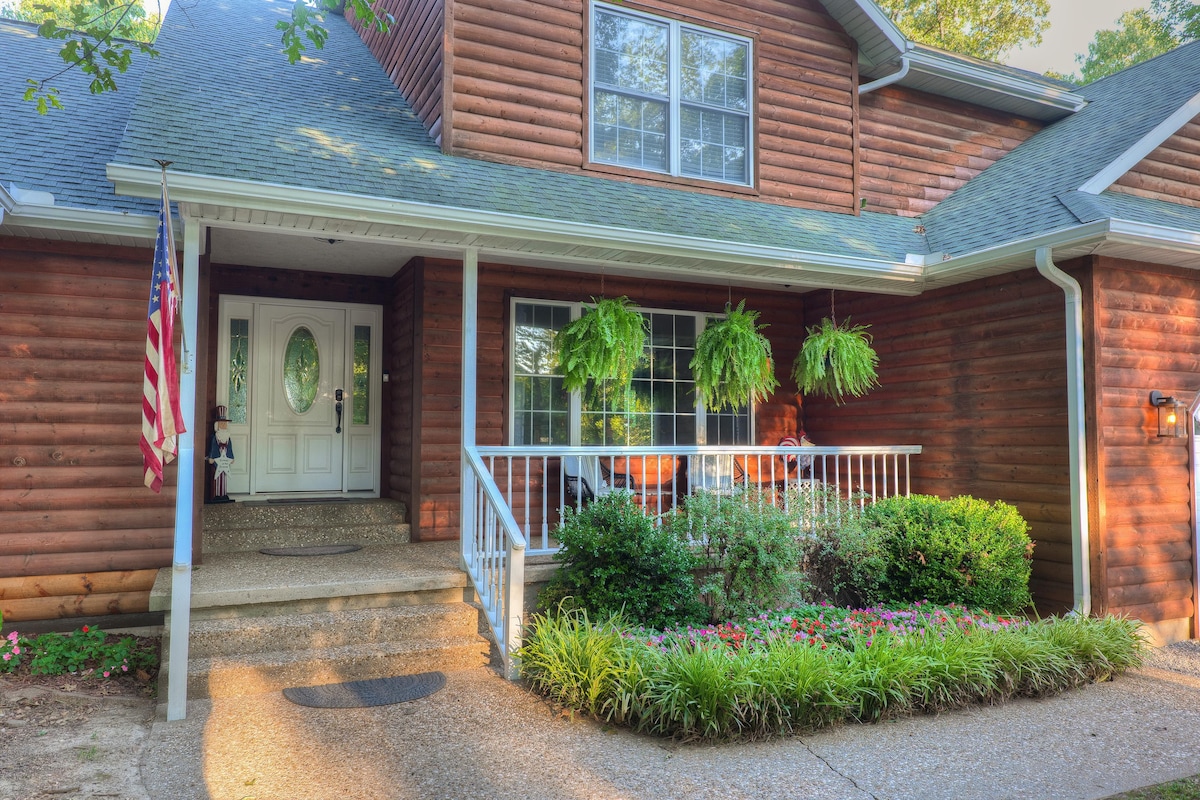 A charming cabin entrance features a covered porch with comfortable seating options. Lush green plants hang from the sides, complementing the wooden exterior. An American flag is displayed on the left, while vibrant flower beds line the walkway leading to the front door.