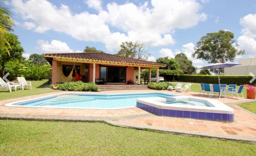 A spacious outdoor area showcases a swimming pool and a relaxing hot tub surrounded by greenery. The house features large glass doors leading to the patio, complemented by comfortable lounge chairs and a shaded umbrella seating area.