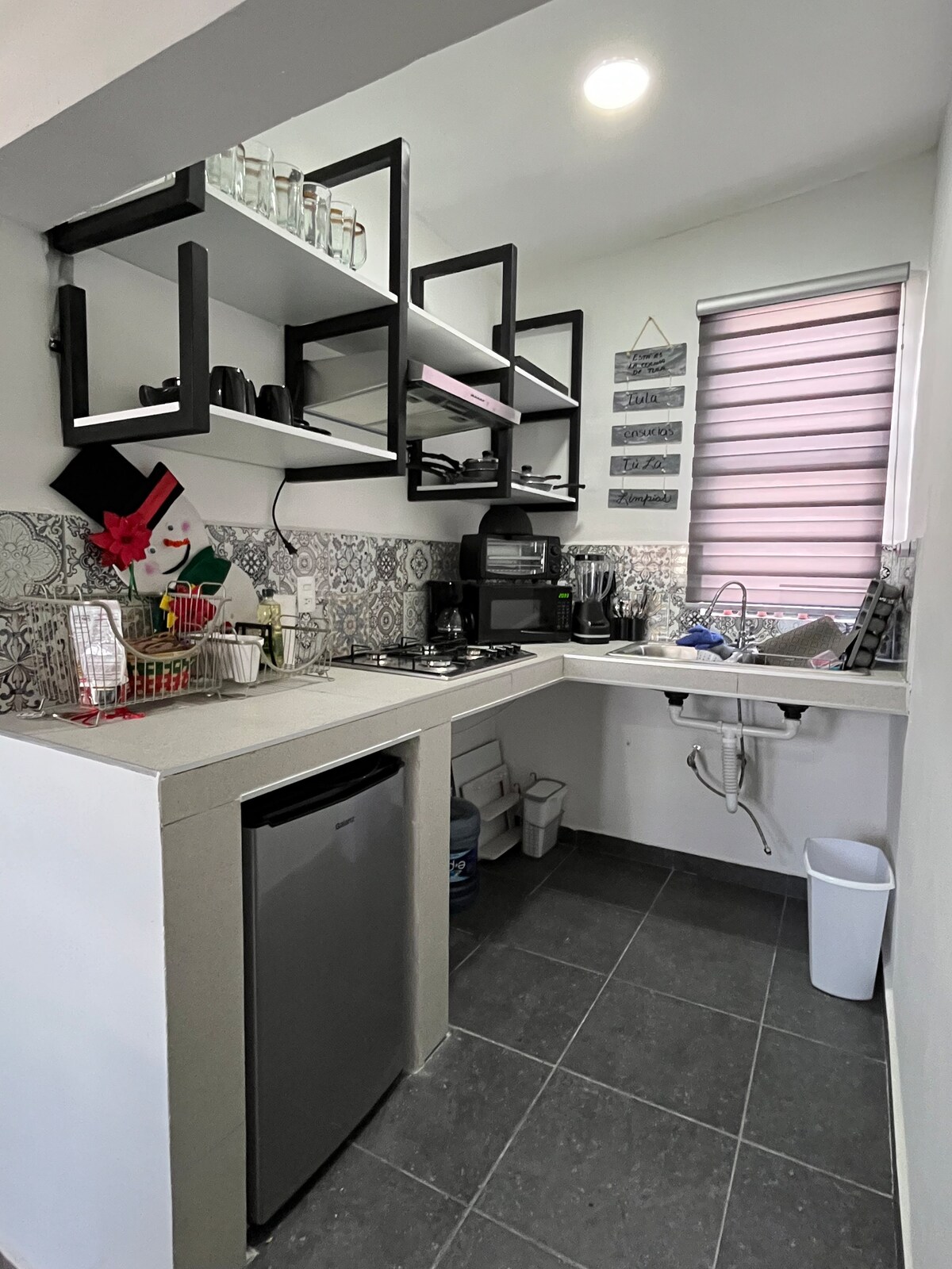 A compact kitchen is equipped with modern appliances and organized shelving for glassware and utensils. A small refrigerator is positioned under the counter, while a gas stove is located on the countertop. Natural light enters through a partially covered window.