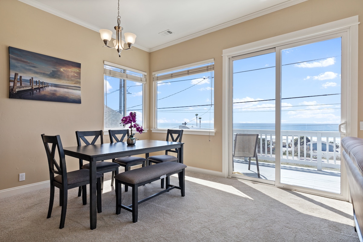 A dining area features a dark wooden table accompanied by six chairs and a long bench, positioned near large windows. Natural light floods the space, revealing stunning views of the ocean. A piece of wall art depicting a pier adds a touch of character.