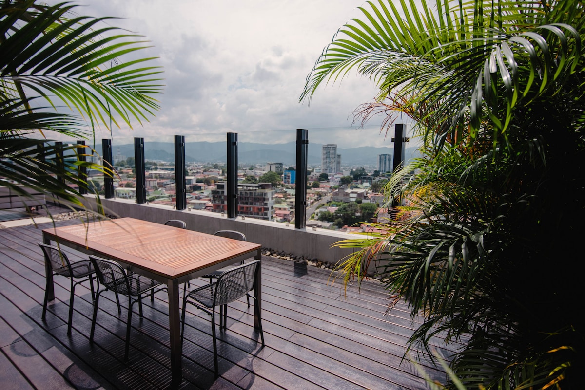 An outdoor terrace features a wooden dining table surrounded by six metal chairs, framed by lush greenery. The view captures a vibrant cityscape and distant mountains under a partly cloudy sky, providing a spacious area ideal for social gatherings.
