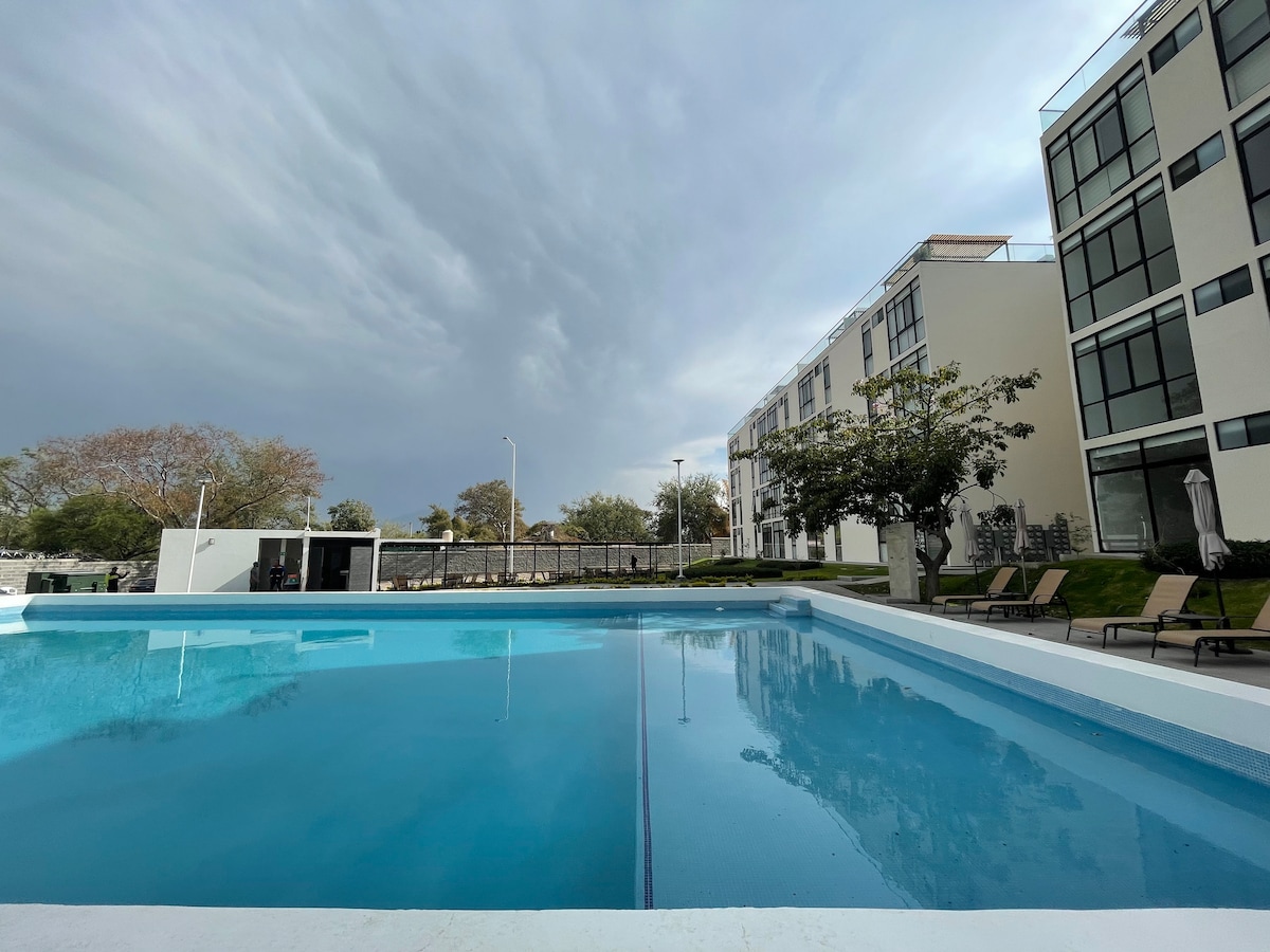 A large swimming pool is shown, reflecting the sky's gray tones. Lounge chairs are positioned around the pool's edge, providing ample space for relaxation. The modern building, with expansive windows, is seen in the background, surrounded by landscaped greenery.