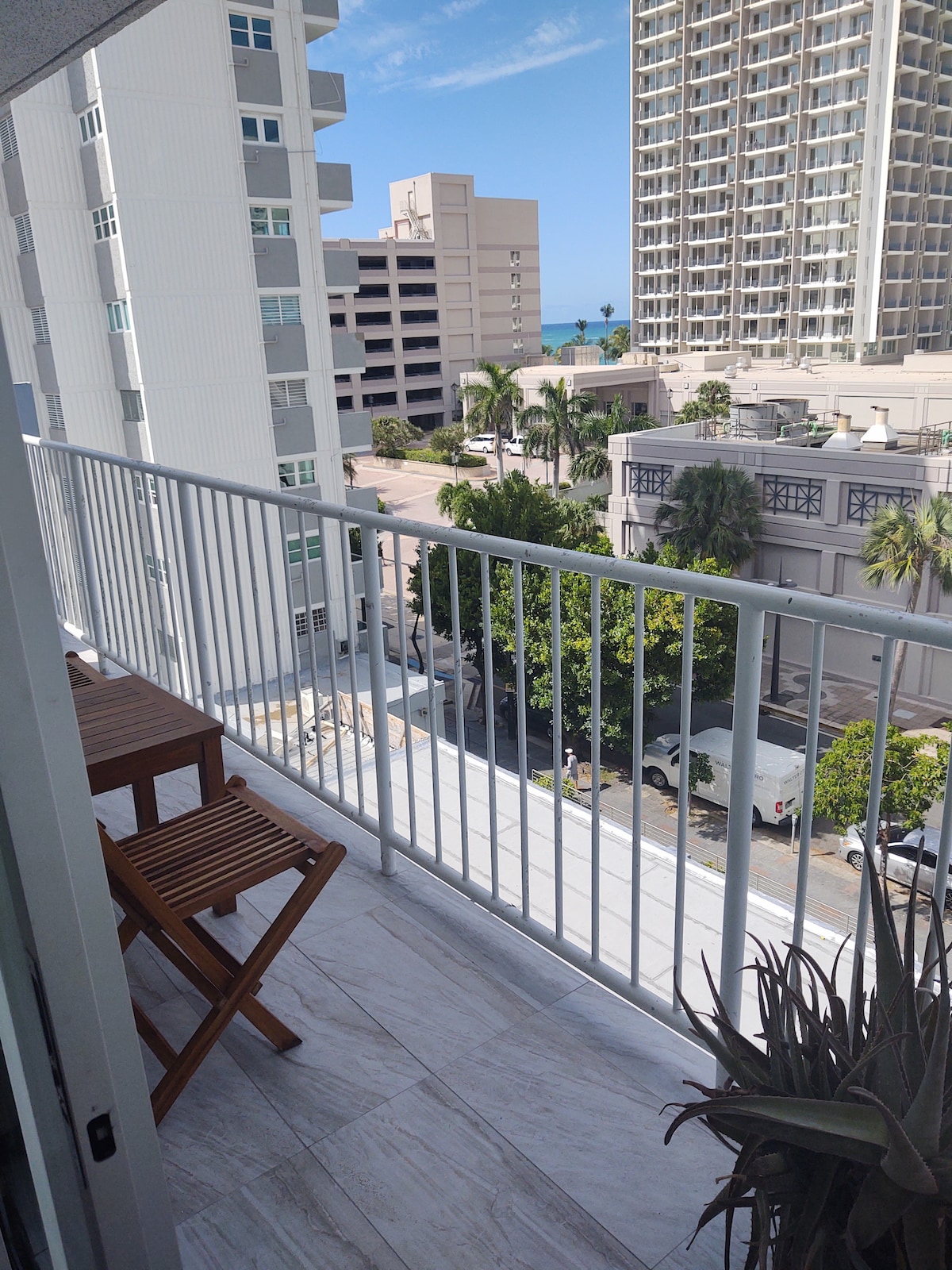 A private balcony is visible, showcasing a small wooden table and two chairs. The railing offers a view of the surrounding buildings and palm trees, with glimpses of the Atlantic Ocean in the distance under a clear blue sky.