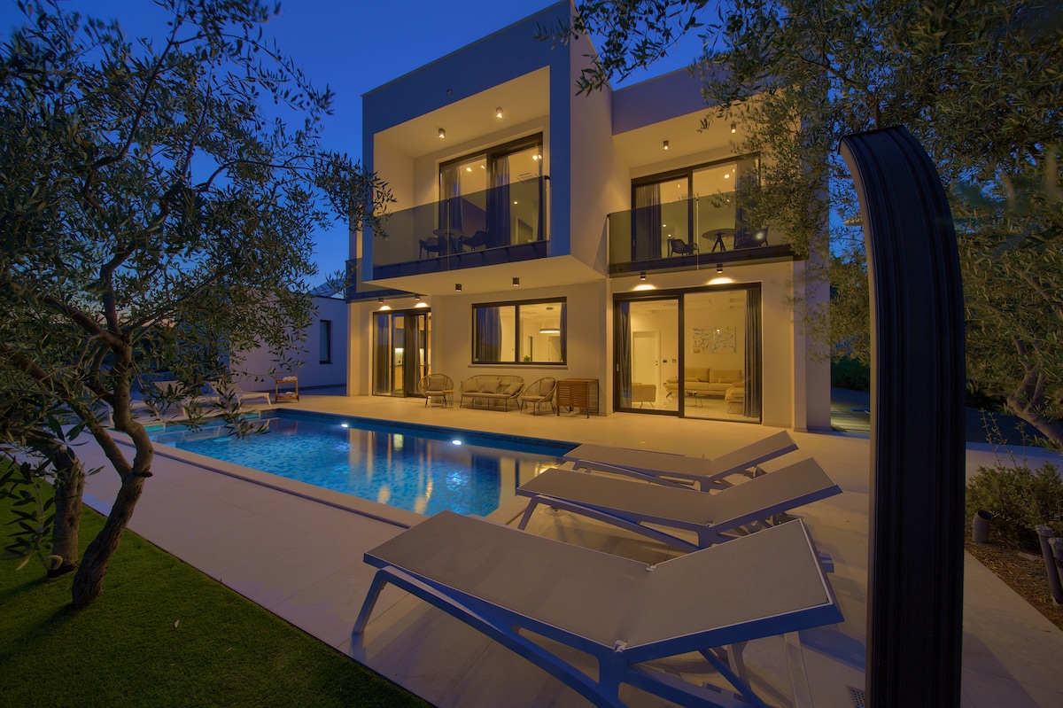 The villa is illuminated at dusk, showcasing a sleek two-story exterior. Sun loungers are positioned by the heated pool, and expansive glass doors provide a view of the interior seating area. The tranquil garden features olive trees, enhancing the serene outdoor ambiance.
