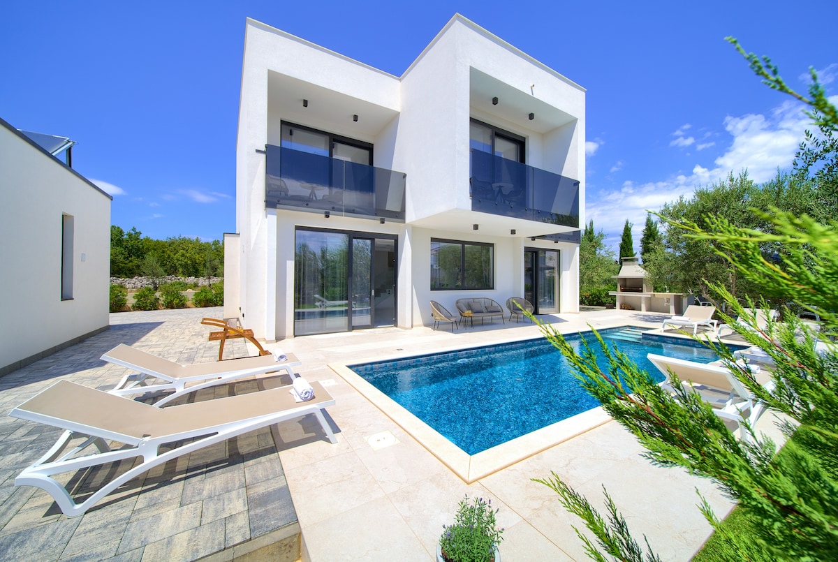 A modern two-story villa is shown with a minimalistic facade and large glass windows. The outdoor area features a heated pool, loungers, and a terrace, surrounded by greenery. Clear skies frame the villa, enhancing the serene environment.