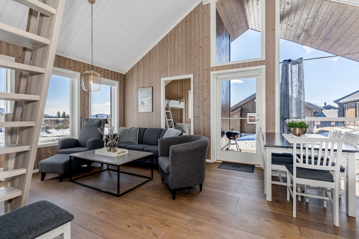 An open living area features large windows that provide ample natural light and views of the outdoors. A comfortable seating arrangement includes a gray sofa and chairs, positioned around a central coffee table. A dining table with white chairs is visible nearby, enhancing the inviting space.