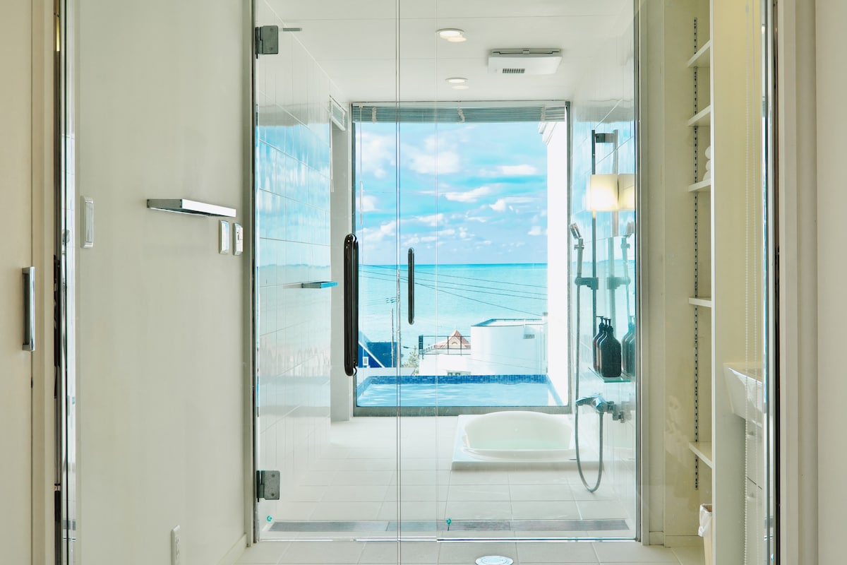 A modern bathroom features large glass doors, providing unobstructed views of the sea. Natural light fills the space, while a freestanding tub sits invitingly next to the glass wall. Essential fixtures are neatly arranged, complementing the overall sleek design.