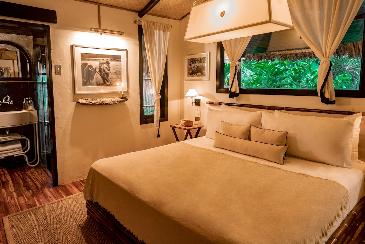 A well-appointed bedroom features a spacious bed dressed in neutral linens, complemented by decorative pillows. Large windows with sheer curtains allow for natural light and views of greenery, while framed black-and-white photographs enhance the decor. A small wooden side table and soft lamp provide additional warmth.