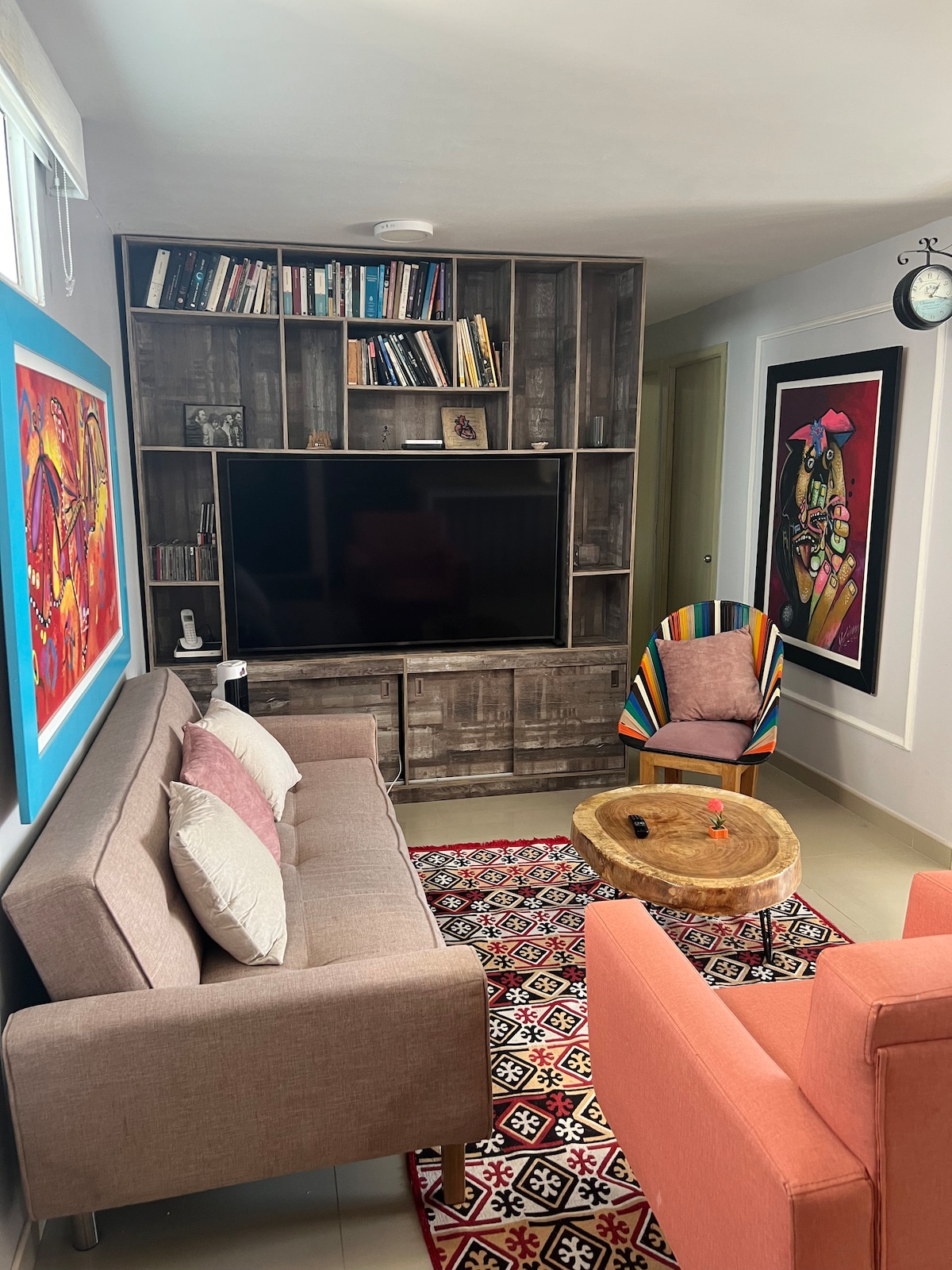 A comfortable living area features a gray sofa and a coral-colored chair, complemented by a coffee table made of natural wood. A contemporary bookshelf holds various books, while vibrant artwork adorns the walls, enhancing the space's design. A large screen television is positioned on a storage unit.