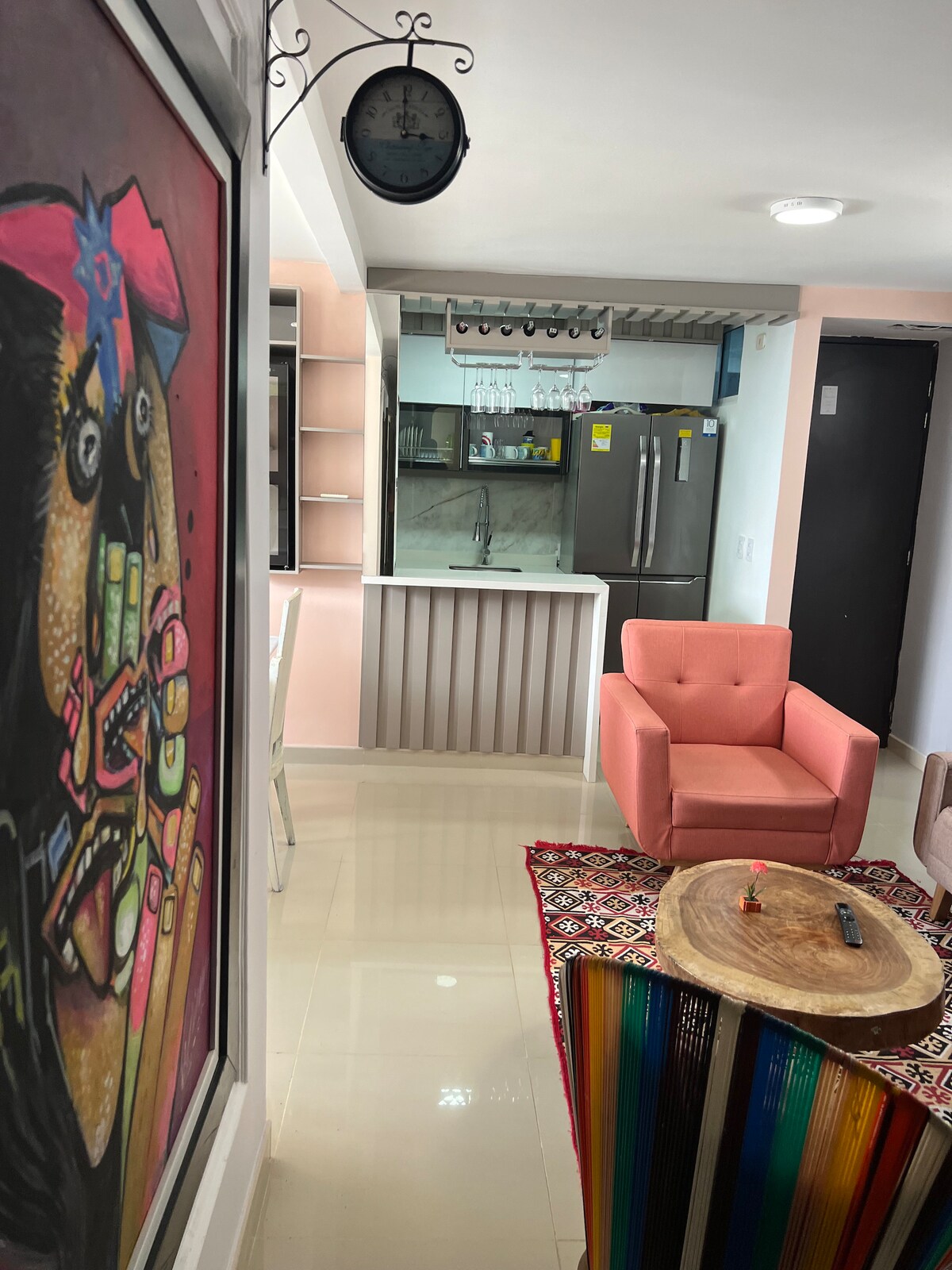 A lively living space is showcased, featuring a vibrant piece of wall art. A pink chair is positioned next to a round wooden table on a colorful rug. The kitchen area is visible in the background, equipped with modern appliances and light cabinetry.