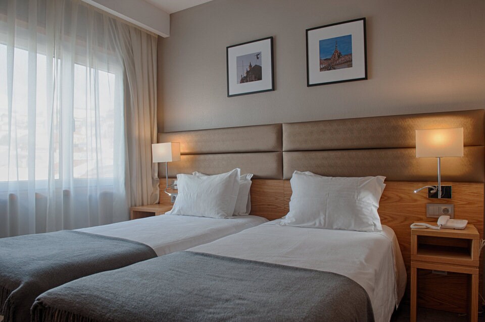 A well-appointed guest room features two spacious beds with crisp white linens and soft grey throws. Natural light filters through a large window, complemented by sheer curtains. Framed artwork adorns the wall above the beds, adding a subtle decorative touch.