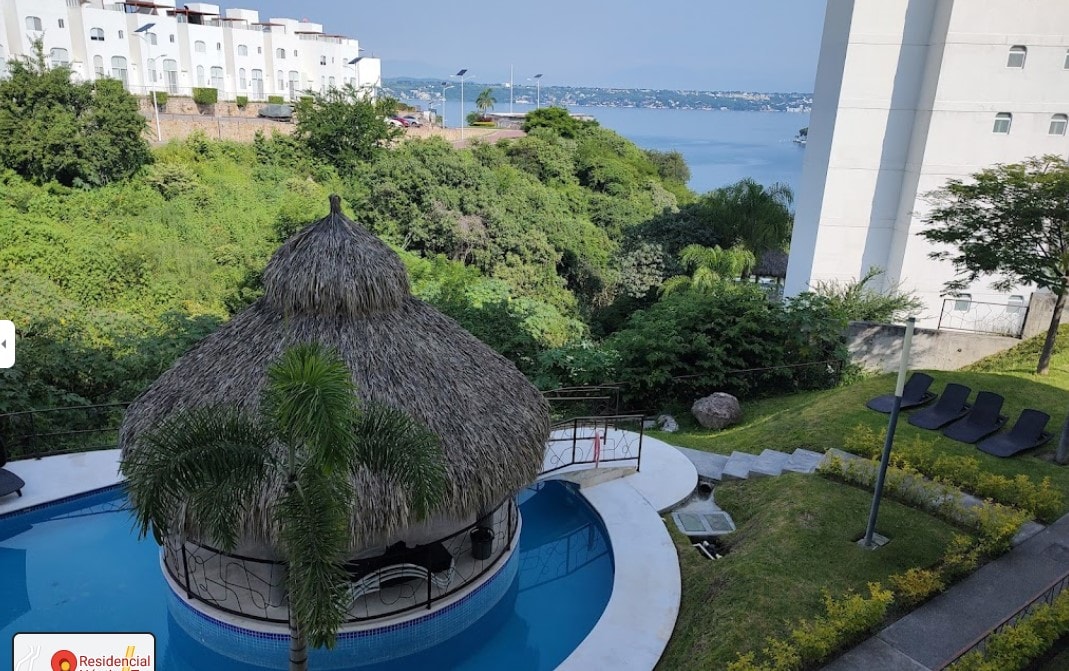 A view of a thatched-roof palapa overlooking a blue pool, surrounded by lush greenery. The serene lake can be seen in the background, creating a peaceful backdrop for outdoor relaxation. Several lounge chairs are positioned near the pool area.