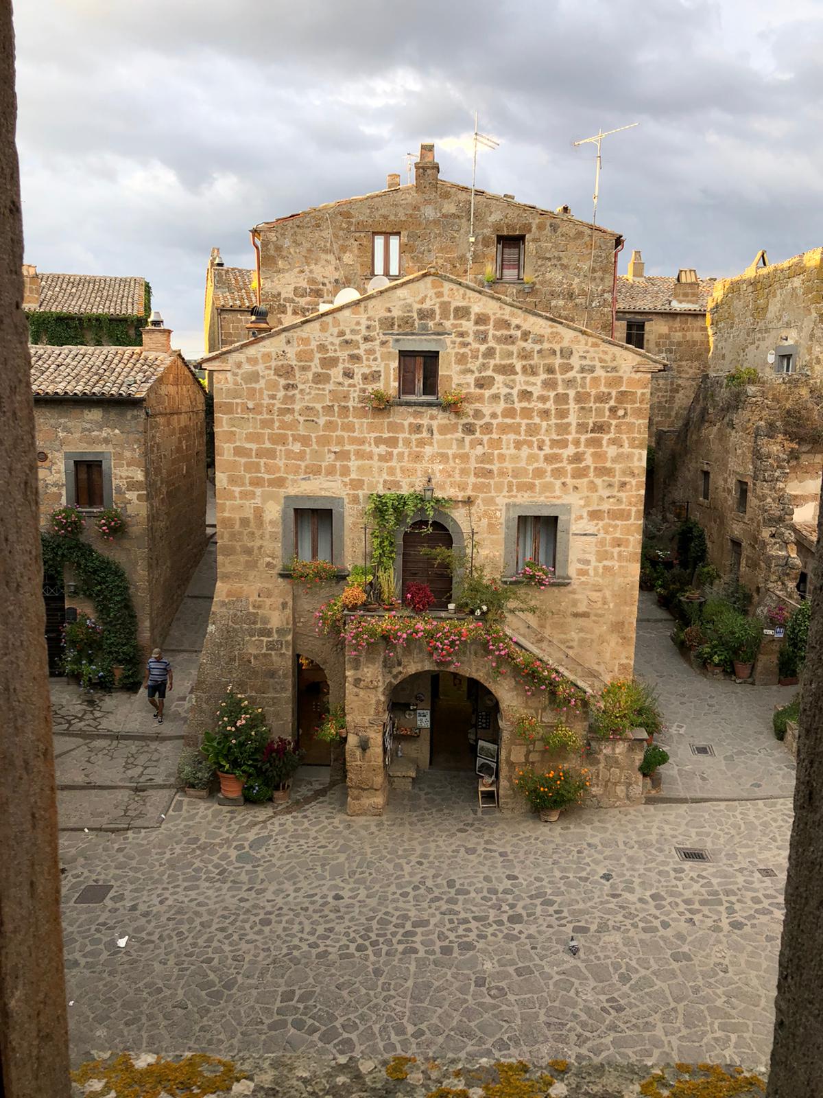 A charming stone building stands at the center of a cobblestone square, adorned with vibrant flowerpots and climbing greenery. The structure features a welcoming archway and several windows, creating an inviting focal point in the quaint village setting. A narrow path leads toward the building.