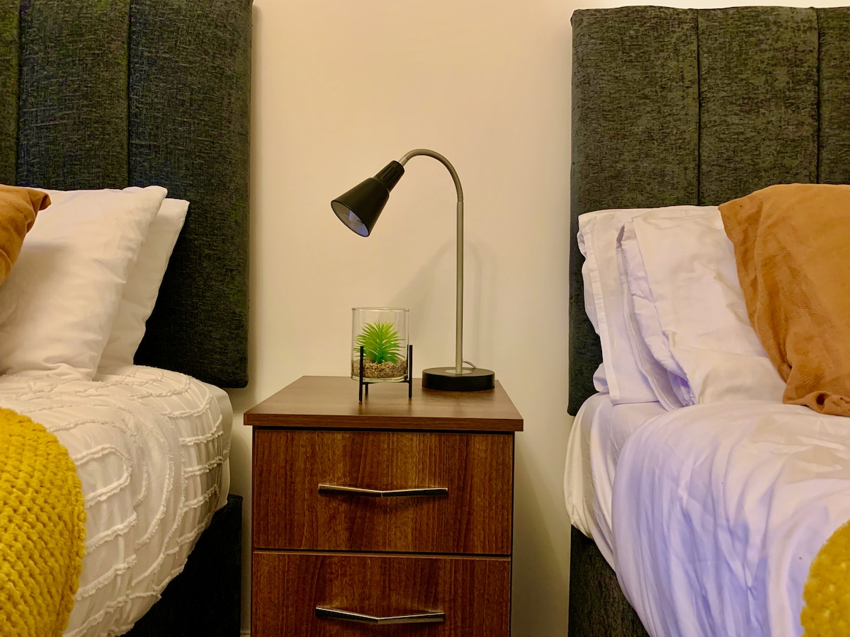 A cozy sleeping area features two beds with plush headboards, positioned on either side of a wooden nightstand. A small lamp rests on the table, beside a decorative plant in glass. Bedding in neutral tones offers a comfortable ambiance.