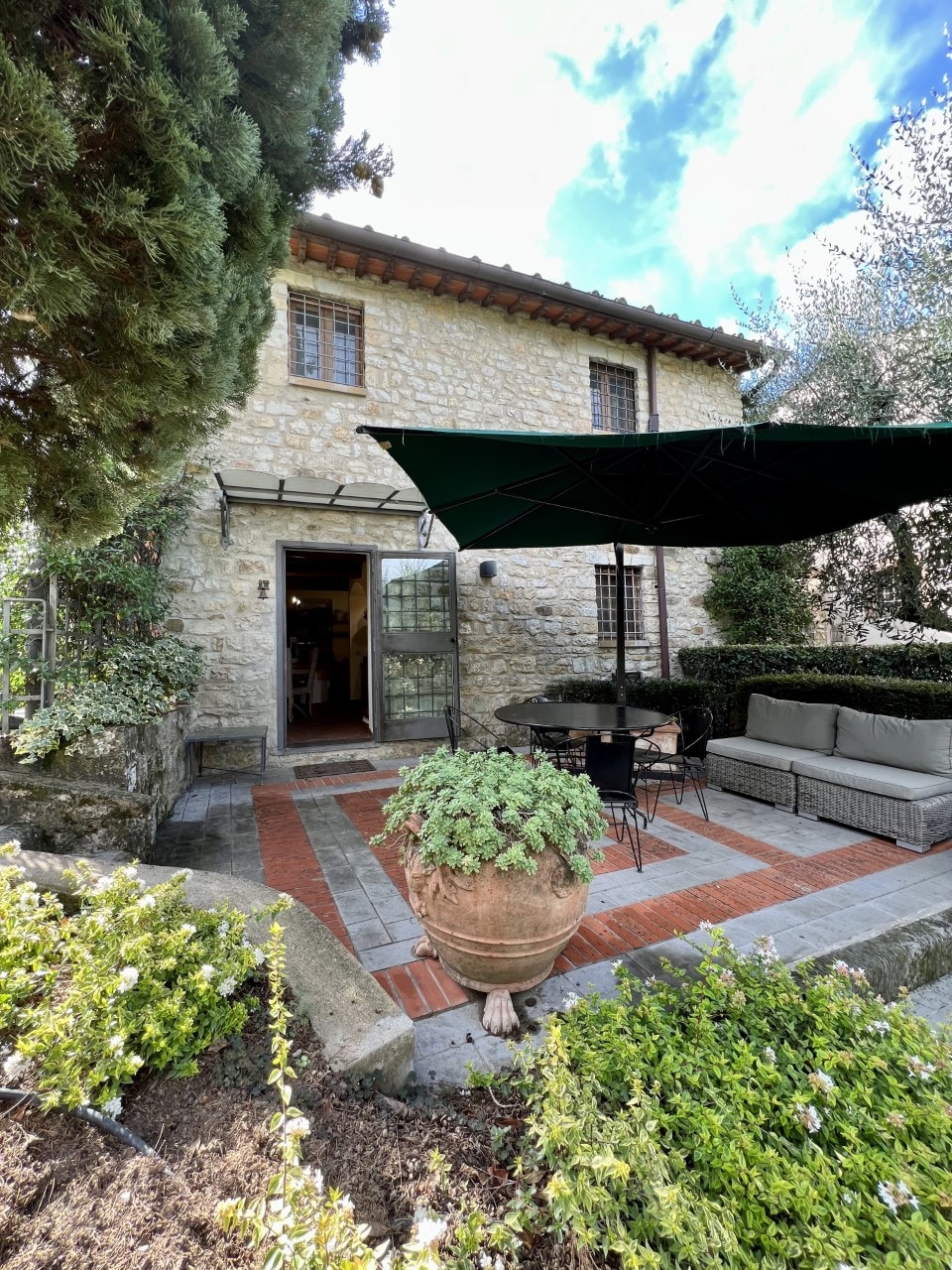 An outdoor terrace features comfortable seating arrangements surrounded by greenery. A large potted plant is positioned at the forefront, while an awning provides shade over the seating area. The rustic stone building is visible in the background, with open glass doors leading inside.