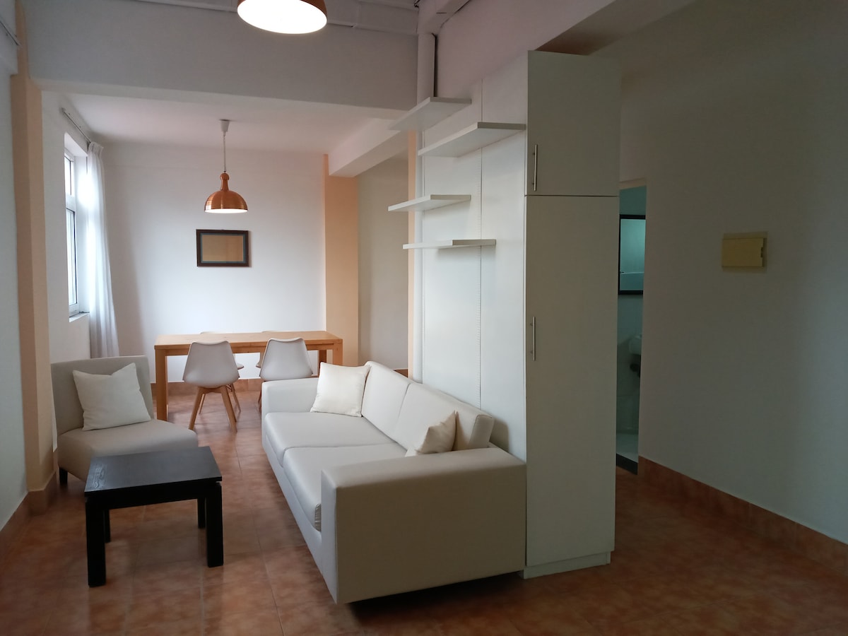 A modern living area features a white sofa and a low black table, creating a minimalist aesthetic. A wooden dining table with chairs is visible in the background. Natural light streams through large windows, illuminating the space. Simple shelving offers additional storage.