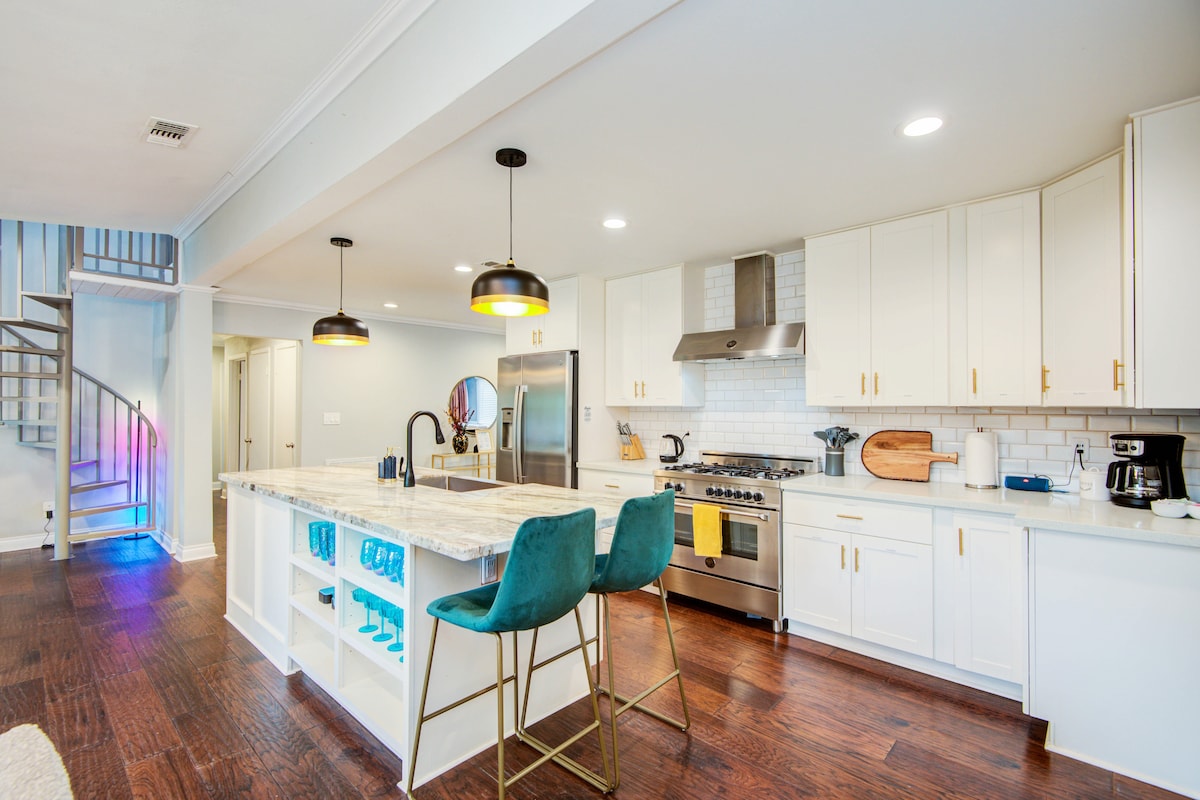 A modern kitchen features white cabinetry and stainless steel appliances. A large island with a marble countertop is accompanied by two teal bar stools. Soft lighting is provided by pendant lights, while a spiral staircase leads to an upper level, creating a bright and inviting space.