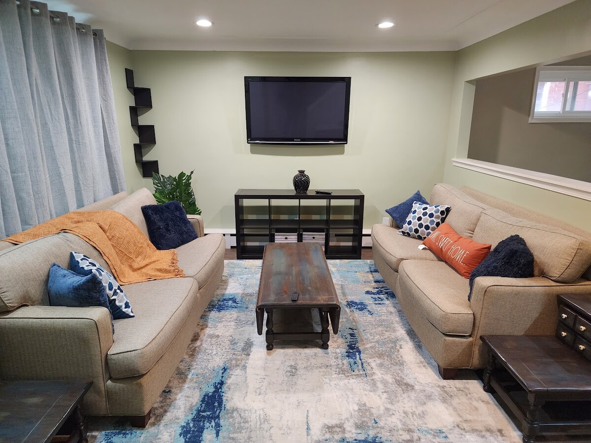 A well-appointed living room features two beige couches adorned with cushions in various shades of blue and orange. A wooden coffee table occupies the center, while a large area rug adds texture underfoot. A television is mounted on the wall, and soft lighting enhances the space.