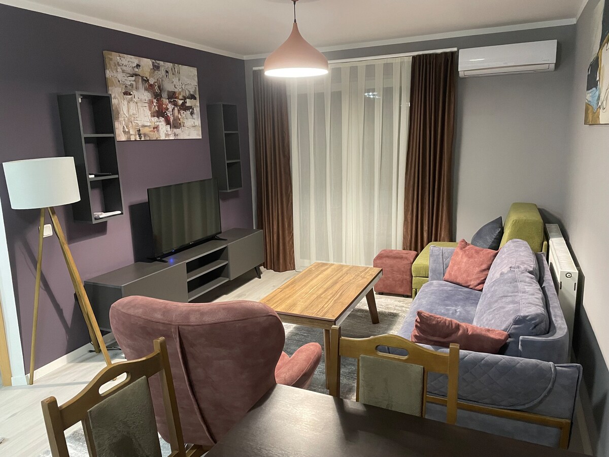 A cozy living room is presented with a mix of colorful seating options, including a spacious sofa and armchairs. A wooden coffee table sits in the center, and a flat-screen TV is mounted on the wall. Natural light filters through sheer curtains, enhancing the inviting ambiance.
