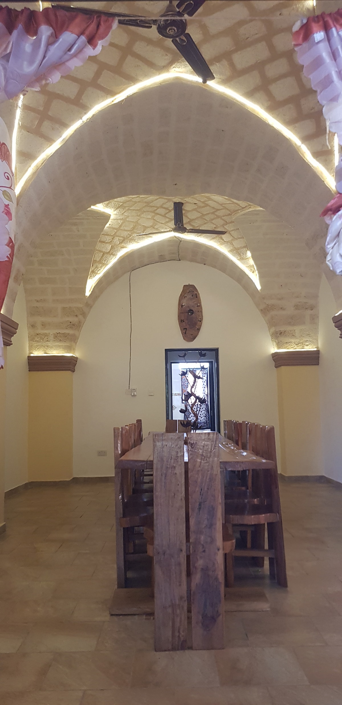 A spacious dining area features a long wooden table surrounded by multiple wooden chairs. The arched ceiling is accented with warm lighting and a unique wall mask is visible through an ornate doorway, creating a welcoming environment.