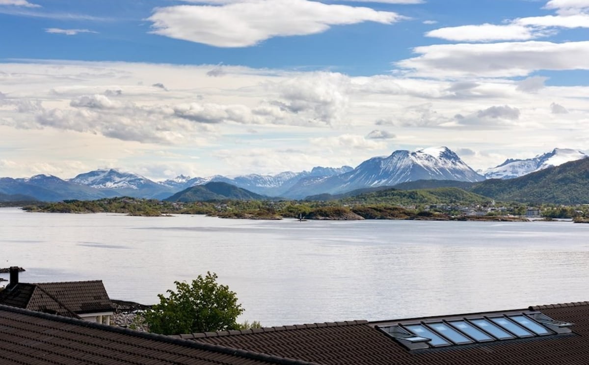 A panoramic view features tranquil water reflecting the sky, with mountains in the background displaying snow-capped peaks. Lush greenery is visible along the shoreline, while a few scattered clouds drift across a bright blue sky.