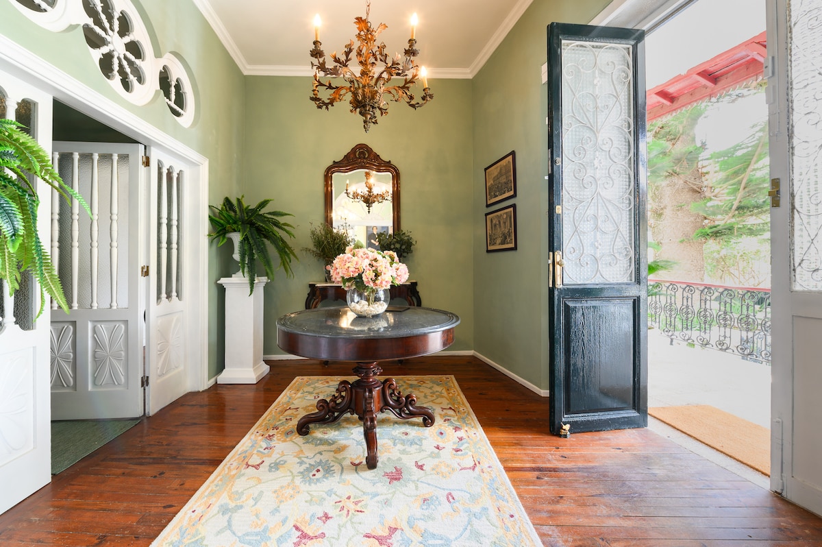 An inviting entryway features elegant wood flooring and a decorative area rug. A round table, adorned with a floral arrangement, complements the ornate chandelier above. The double doors open to the outdoor space, framed by detailed woodwork and greenery.