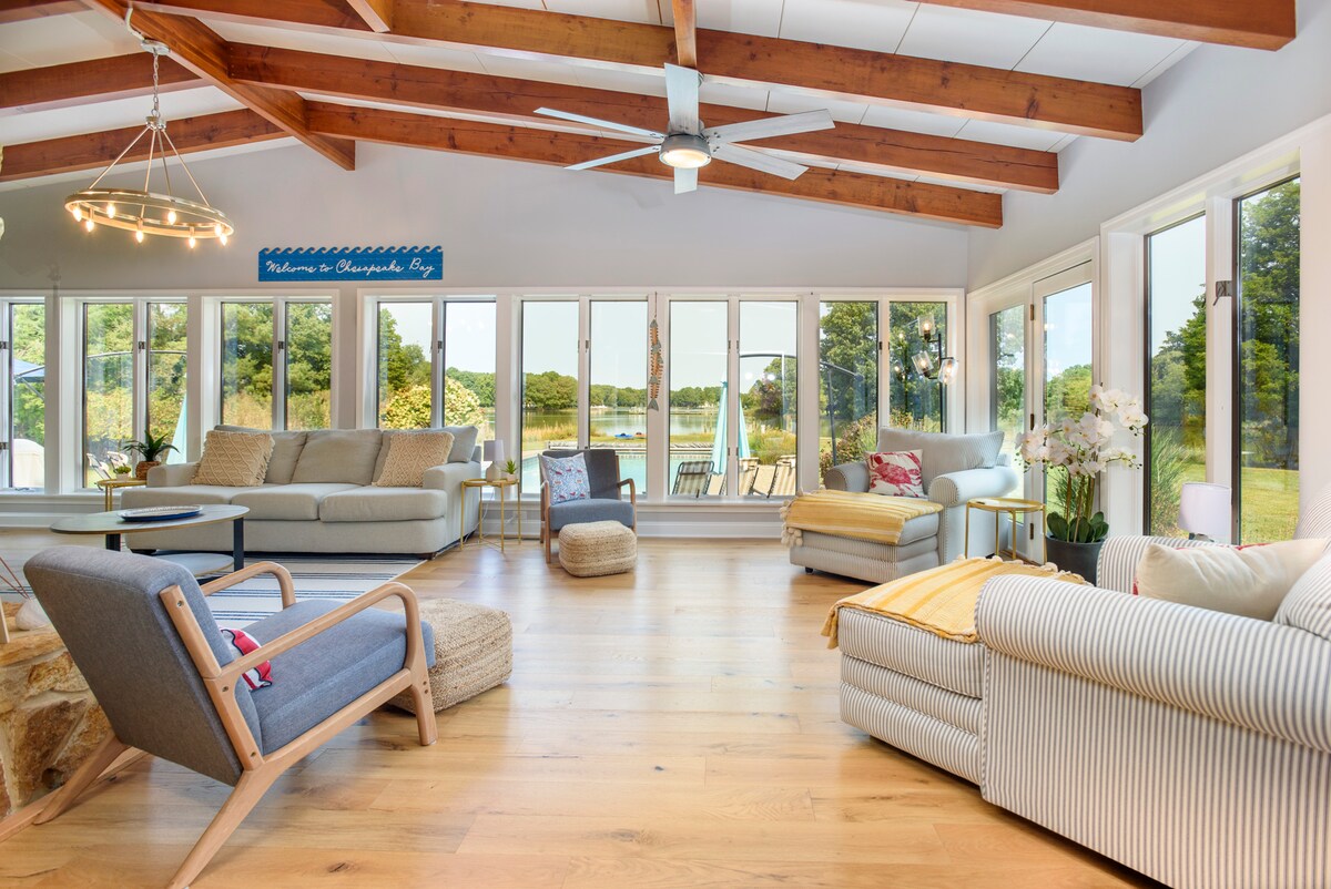 A spacious living area features large windows allowing natural light to fill the room. The ceiling showcases exposed wooden beams, while a mix of comfortable seating options, including sofas and armchairs, is arranged thoughtfully. Views of the waterfront can be seen from this inviting space.