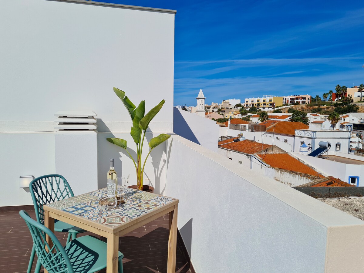 A private terrace is shown, featuring a small table with two chairs, surrounded by a vibrant plant. The view highlights rooftops of Ferragudo, with a clear blue sky above, creating a peaceful outdoor space for relaxation.