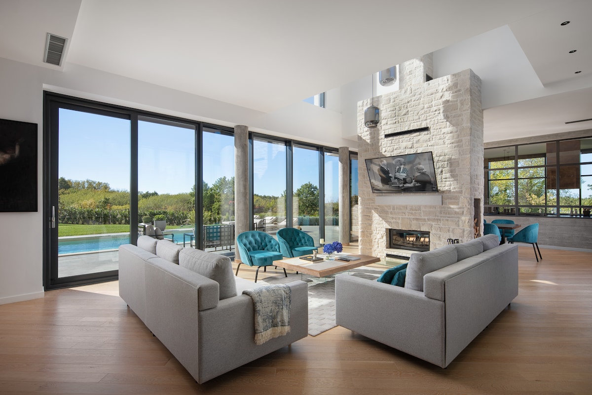 A light-filled living room features modern furniture, including two grey sofas and turquoise accent chairs. A coffee table is centered on a soft rug, with a stone fireplace serving as a focal point. Floor-to-ceiling windows offer views of a serene outdoor pool and greenery.
