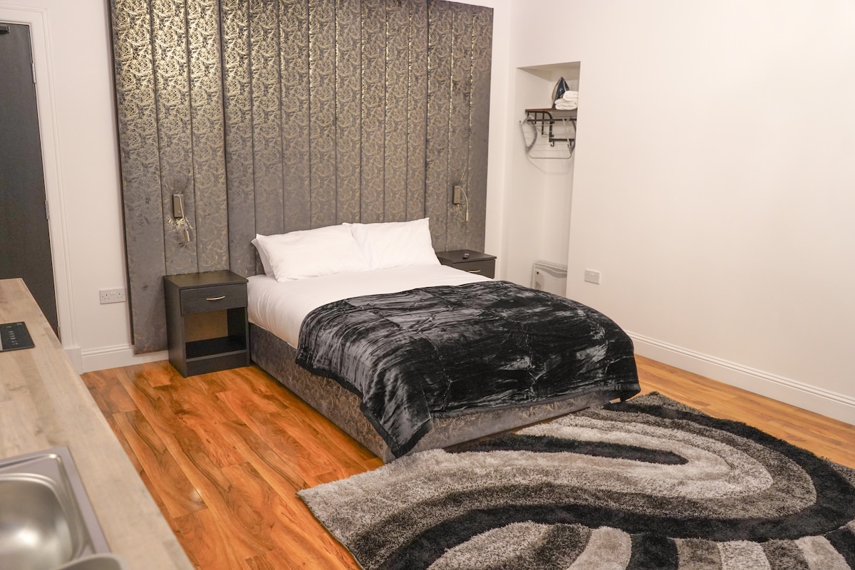 A spacious sleeping area features a plush bed with a textured headboard. Dark wood nightstands are positioned on either side. A soft, patterned rug complements the hardwood floor. A small kitchenette and storage area are visible in the background, enhancing the studio's functionality.