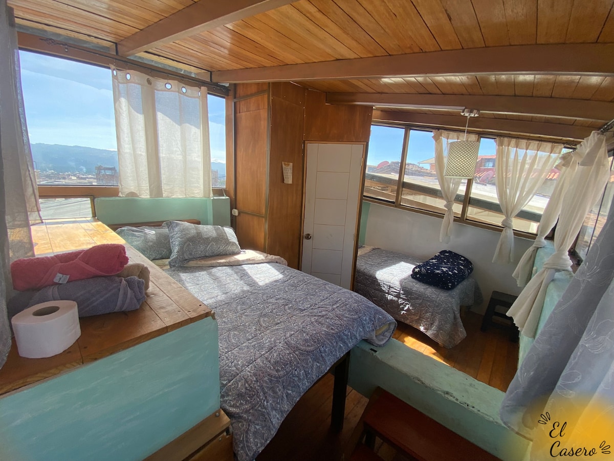 A cozy room with wooden walls features two spacious beds, one with a textured blue and white blanket. Large windows allow natural light to flood the space, while sheer curtains frame the views. A small table is visible, enhancing the room’s functionality.