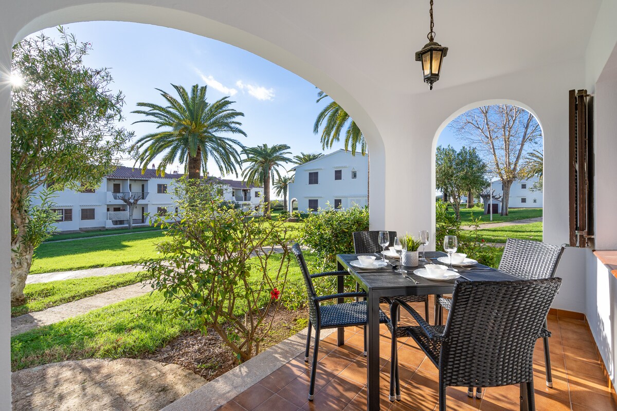 An outdoor terrace is presented, featuring a dining table with four chairs. Surrounding greenery and palm trees create a serene environment. The view includes residential buildings in the background, visible through the arches of the terrace.