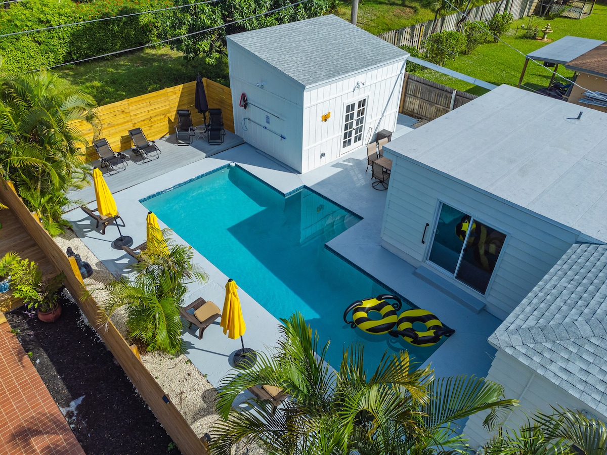 The aerial view displays a heated pool surrounded by lounge chairs and yellow umbrellas. The pool area features two colorful pool floats. Enclosed by lush greenery, the exterior space offers privacy and a relaxing environment for guests.
